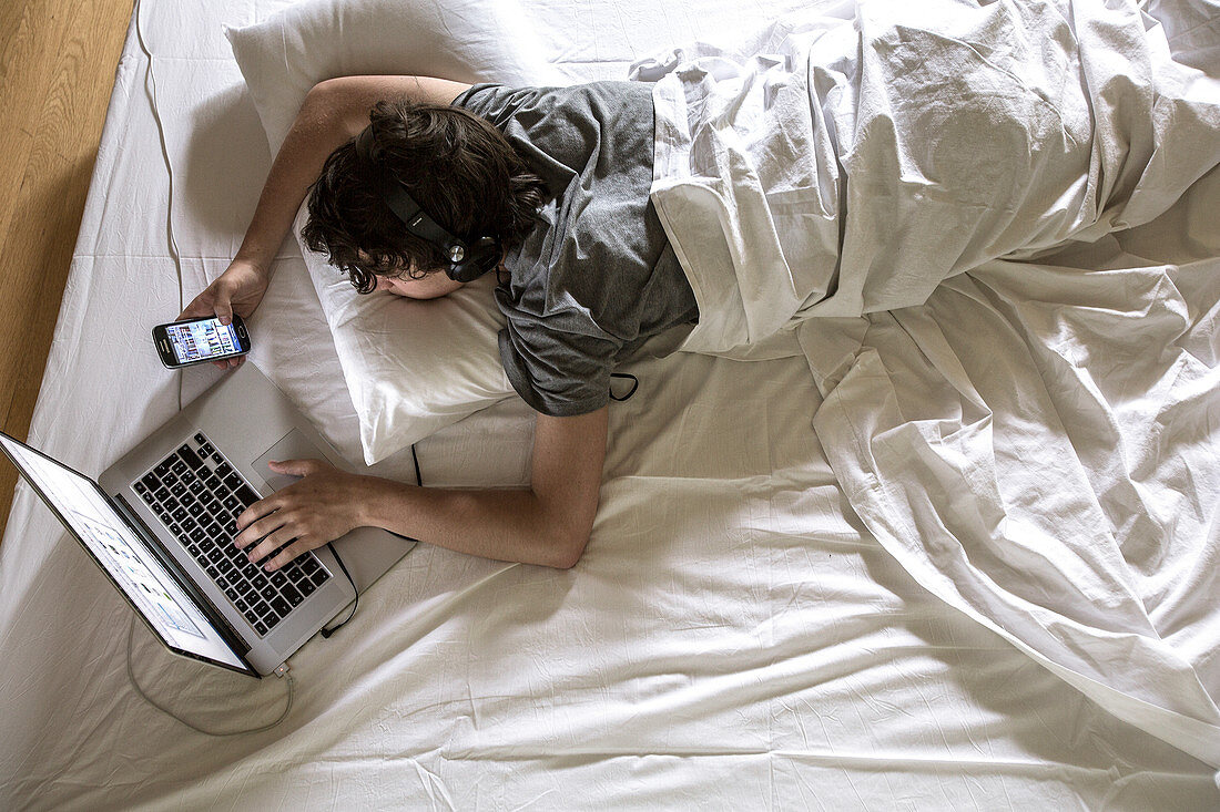 Man lying in bed using laptop and phone
