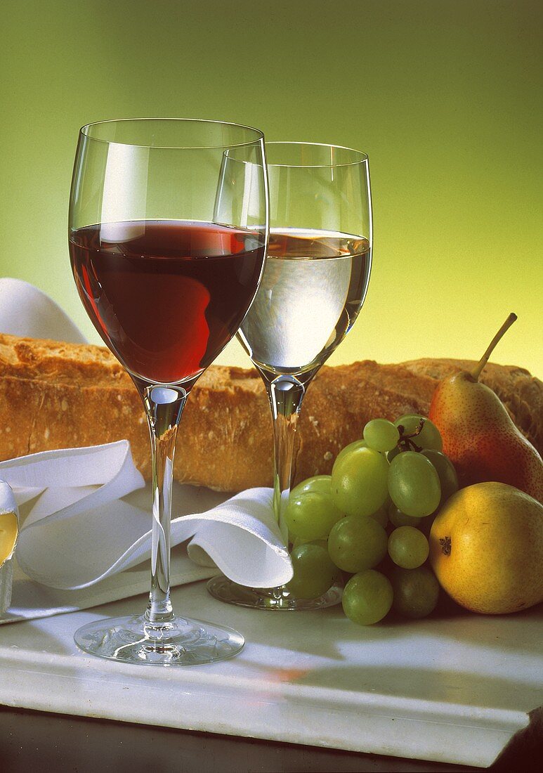 Zwei Gläser mit Weiß- und Rotwein vor Obst und einem Baguette