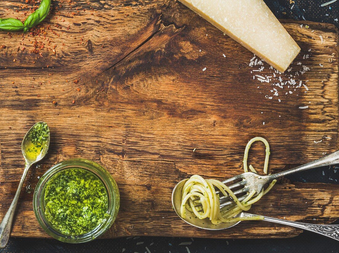 Jar with pesto sauce, spoon and fork with cooked spaghetti, fresh basil leaves and Parmesan cheese