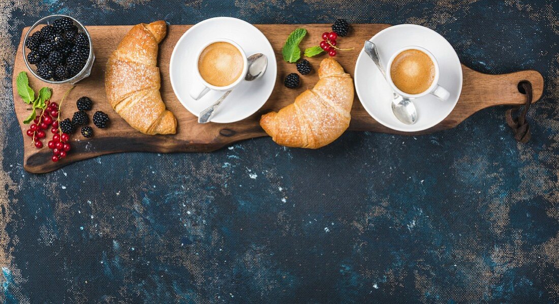 Freshly baked croissants with garden berries and coffee cups served on rustic wooden board