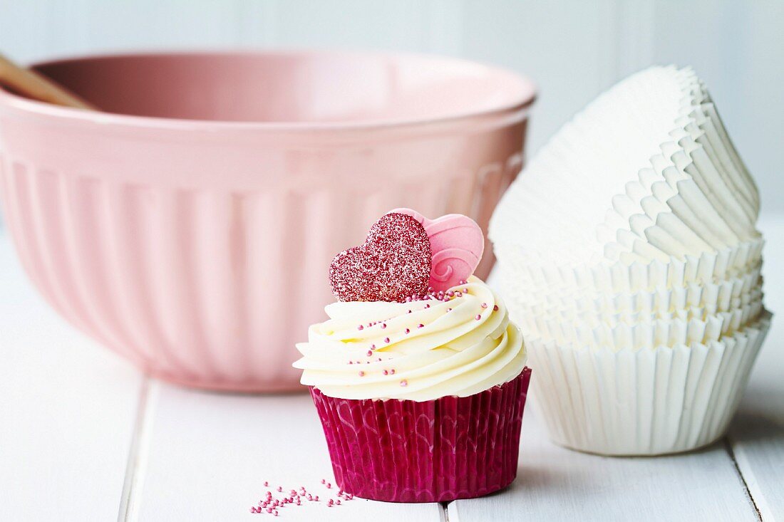 Cupcake mit Herzdekor vor Papierbackförmchen und Schüssel