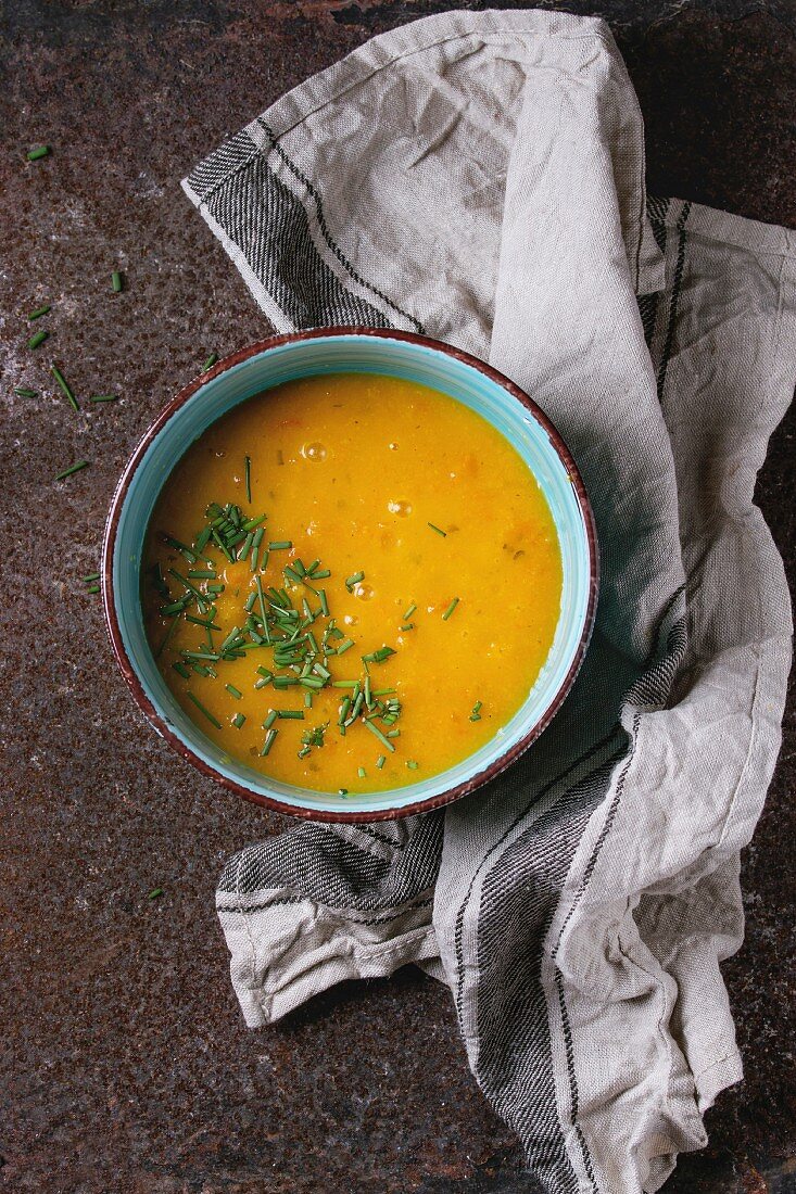 Blue ceramic bowl of carrot soup, served with onion chives and kitchen towel over old rusty iron background