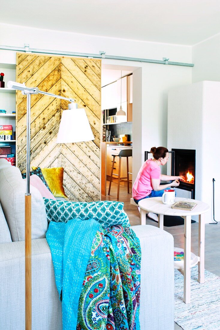 Woman lighting fire in stove in living area with rustic sliding door leading to kitchen