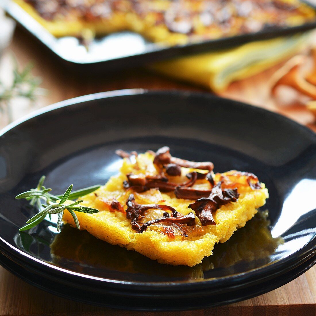 A slice of polenta cake with chanterelle mushrooms and onions on a plate in front of a baking tray