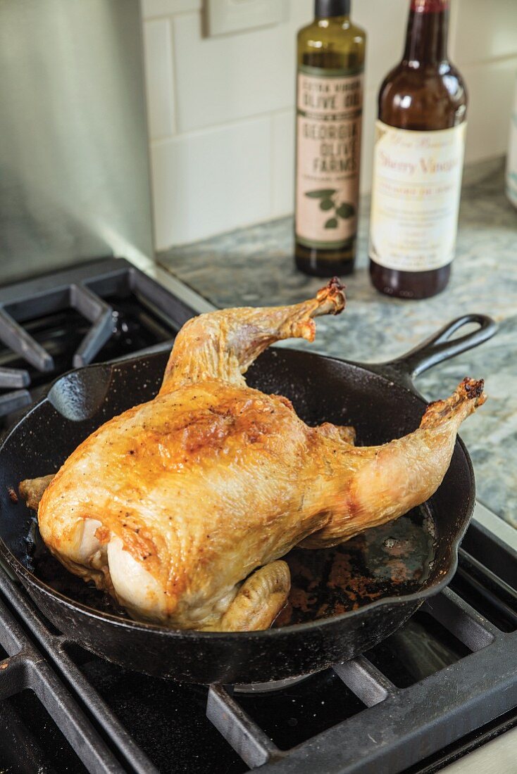 Roast chicken in a cast iron pan on a stove with bottles of oil in the background