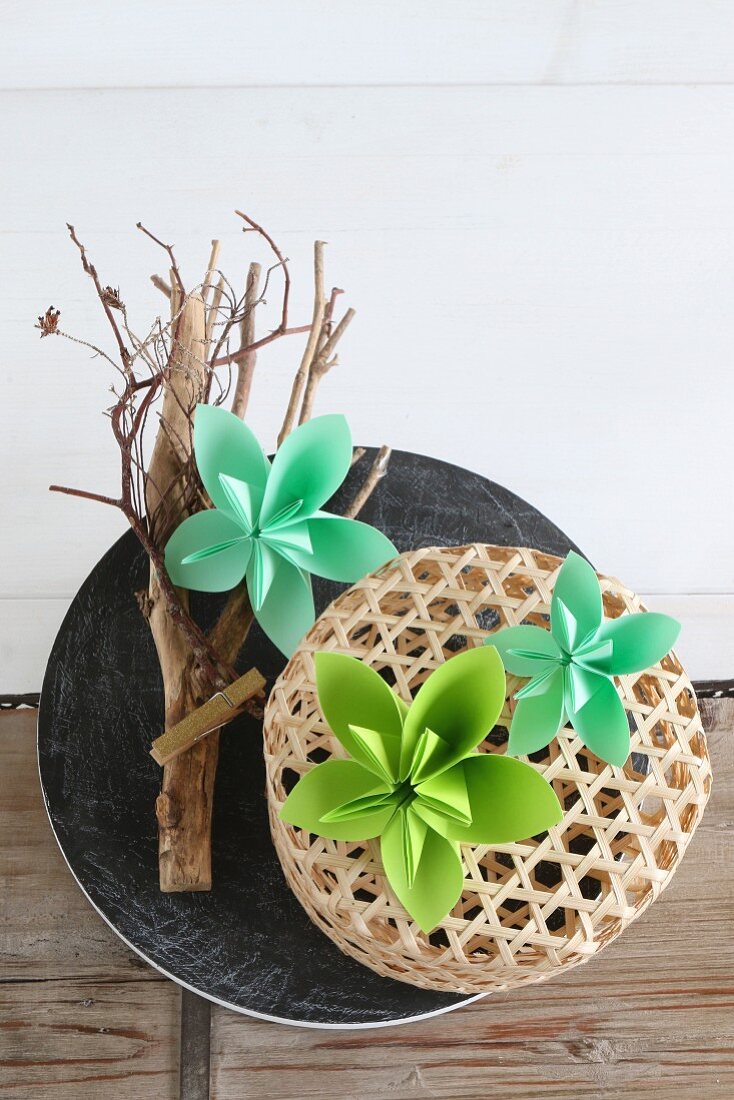 Grüne Origami-Blüten auf einem Ast und Bastkörbchen