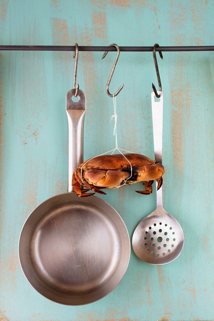 A crab hanging from a hook between a saucepan and a draining spoon