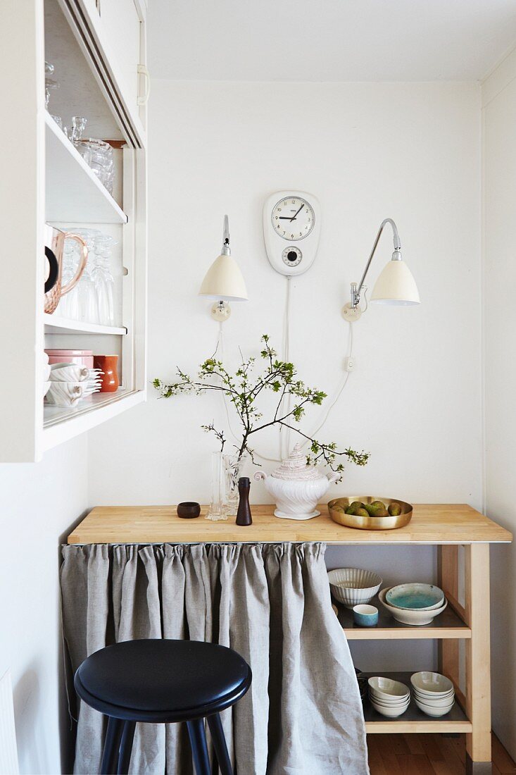 Wooden table with crockery on shelves below behind pale grey curtain under wall lamps and retro wall clock
