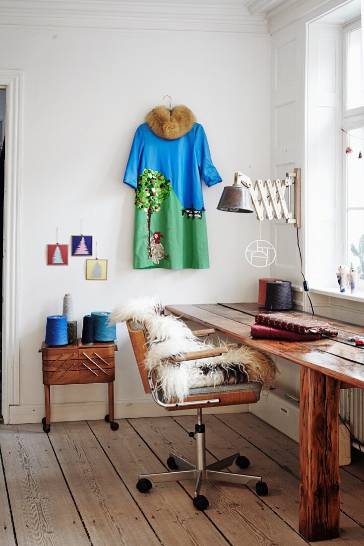 Rustic wooden table, sheepskin on retro office chair next to reels of thread on sewing box below dress hung on wall