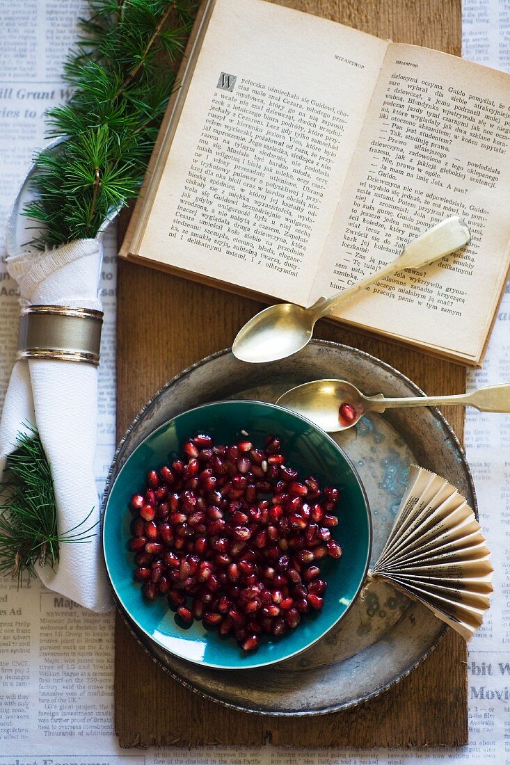 Pomegranate seeds in a bowl with a silver spoon and an open book