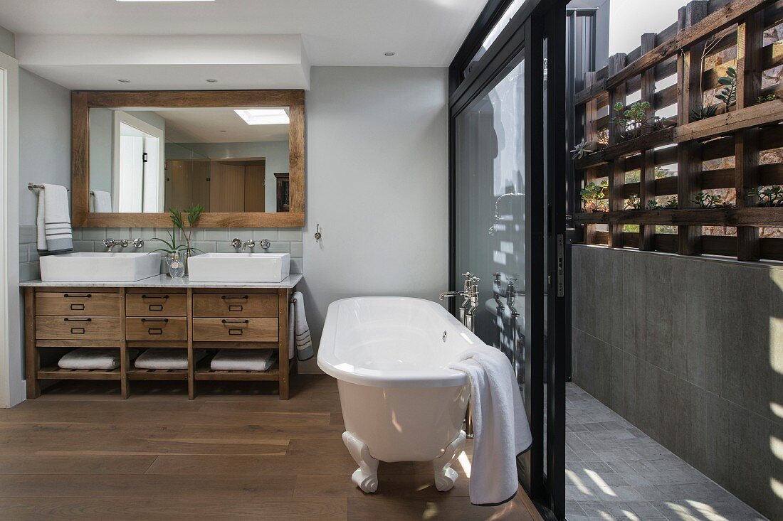 White, free-standing bathtub in front of open sliding doors leading to narrow courtyard with rustic wooden screen