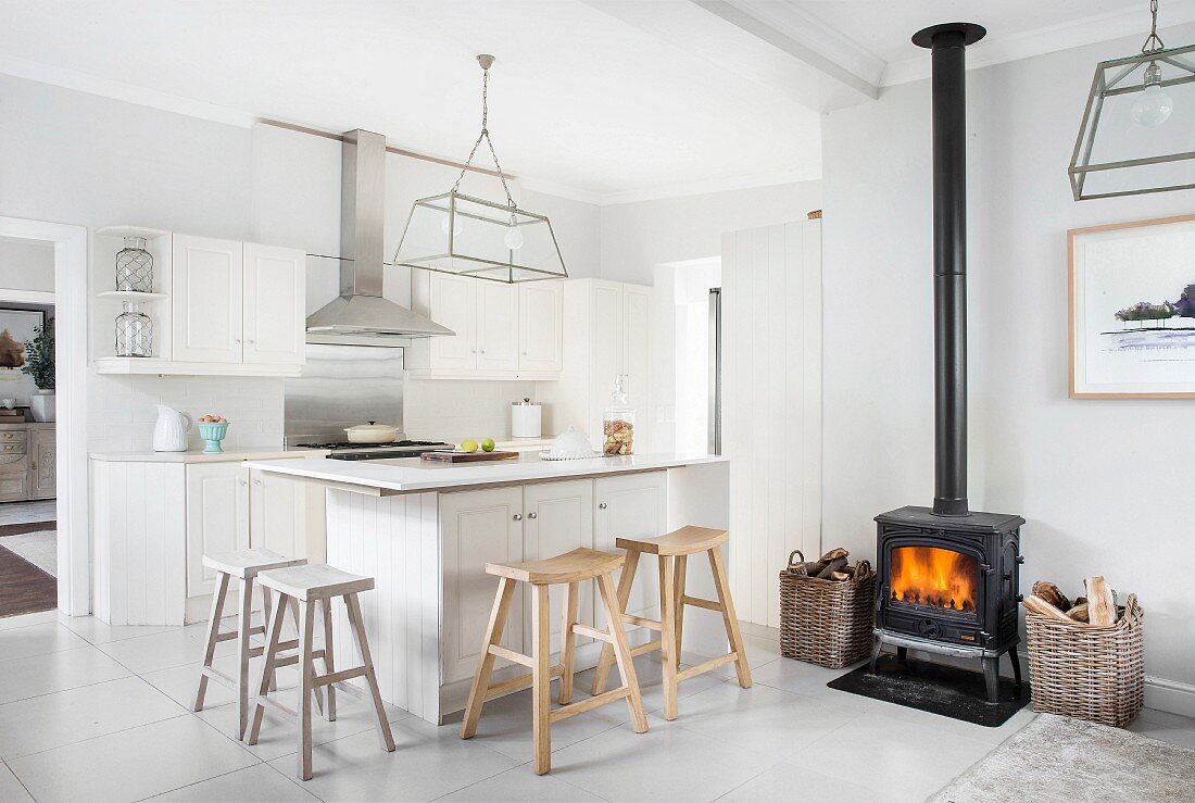 Black log-burning stove and rustic wooden bar stools at counter in white, open-plan kitchen