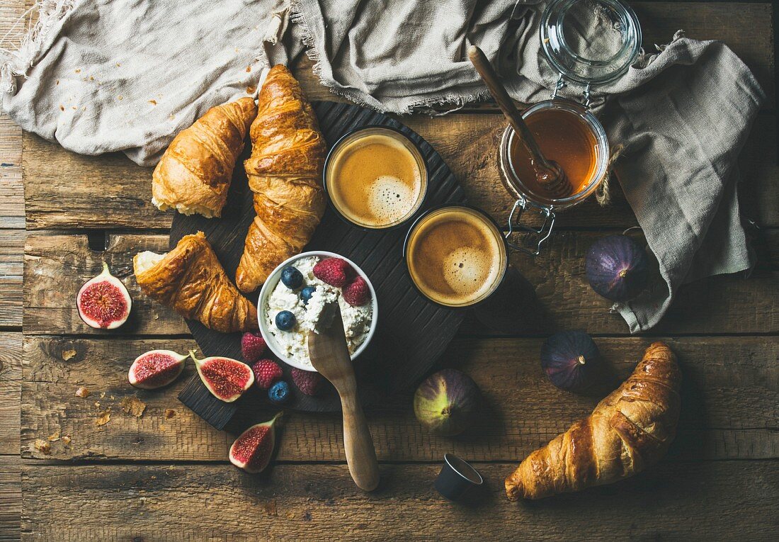 Frühstück mit Croissants, Ricotta, Feigen, Beeren, Honig und Espresso auf Holzuntergrund