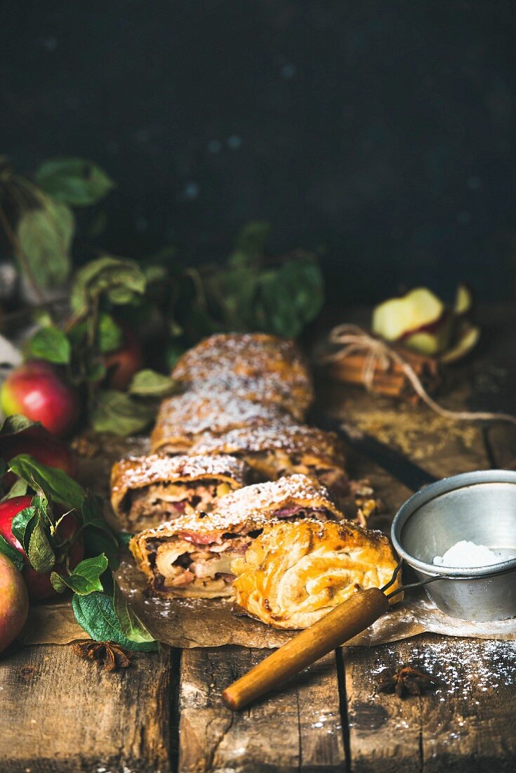 Sliced apple strudel with fresh apples, icing sugar and spices on a rustic wooden table