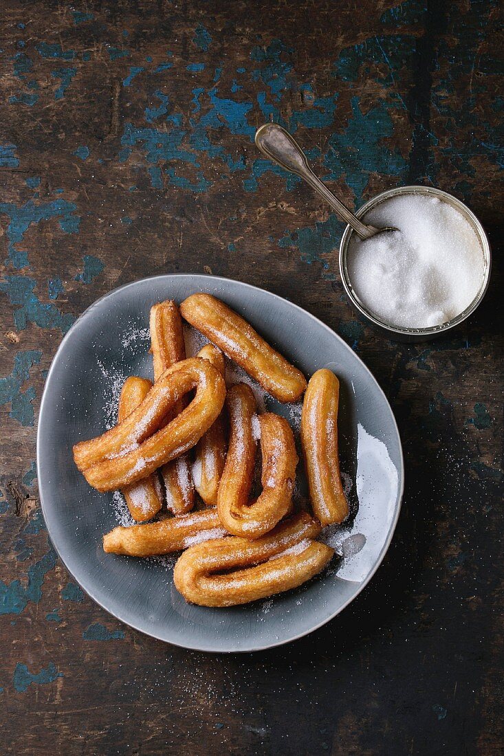 Traditional spanish treat churros with granulated sugar, served in gray oval plate with tin can of sugar