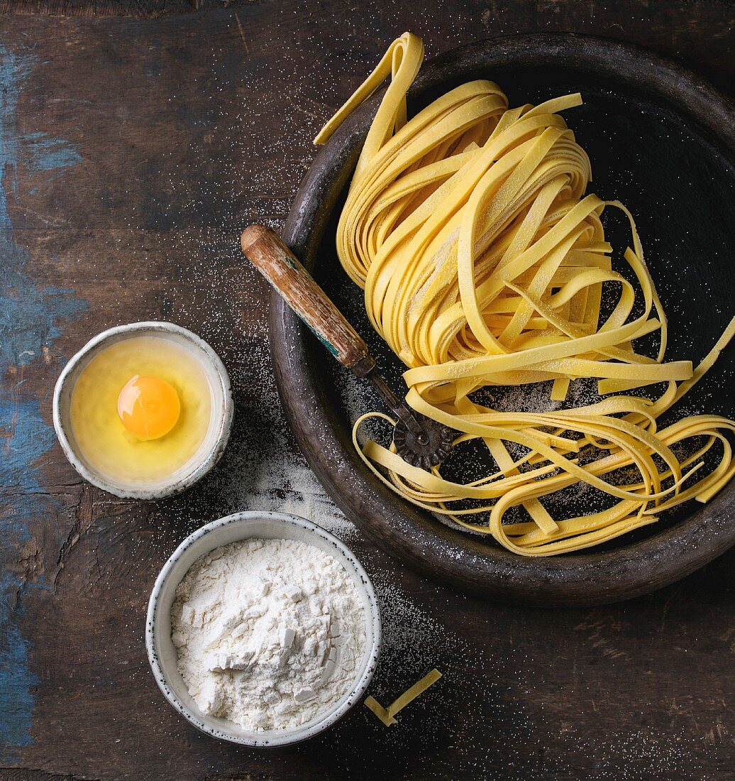 Raw uncooked homemade italian pasta tagliatelle with pasta cutter, bowls with white flour and broken egg