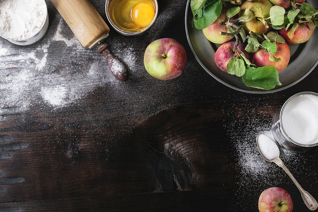 Ingredients for making apple cake: flour, sugar, a whisked egg, fresh apples, sugar and a rolling pin