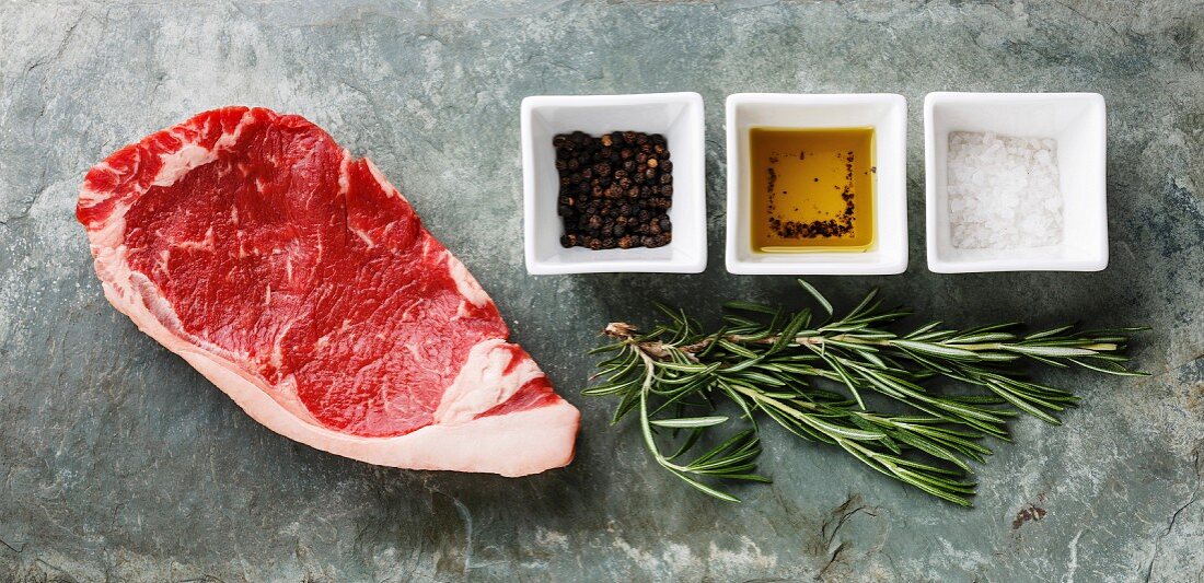 Raw fresh meat steak Striploin with rosemary, cooking oil, salt and pepper on stone slate background
