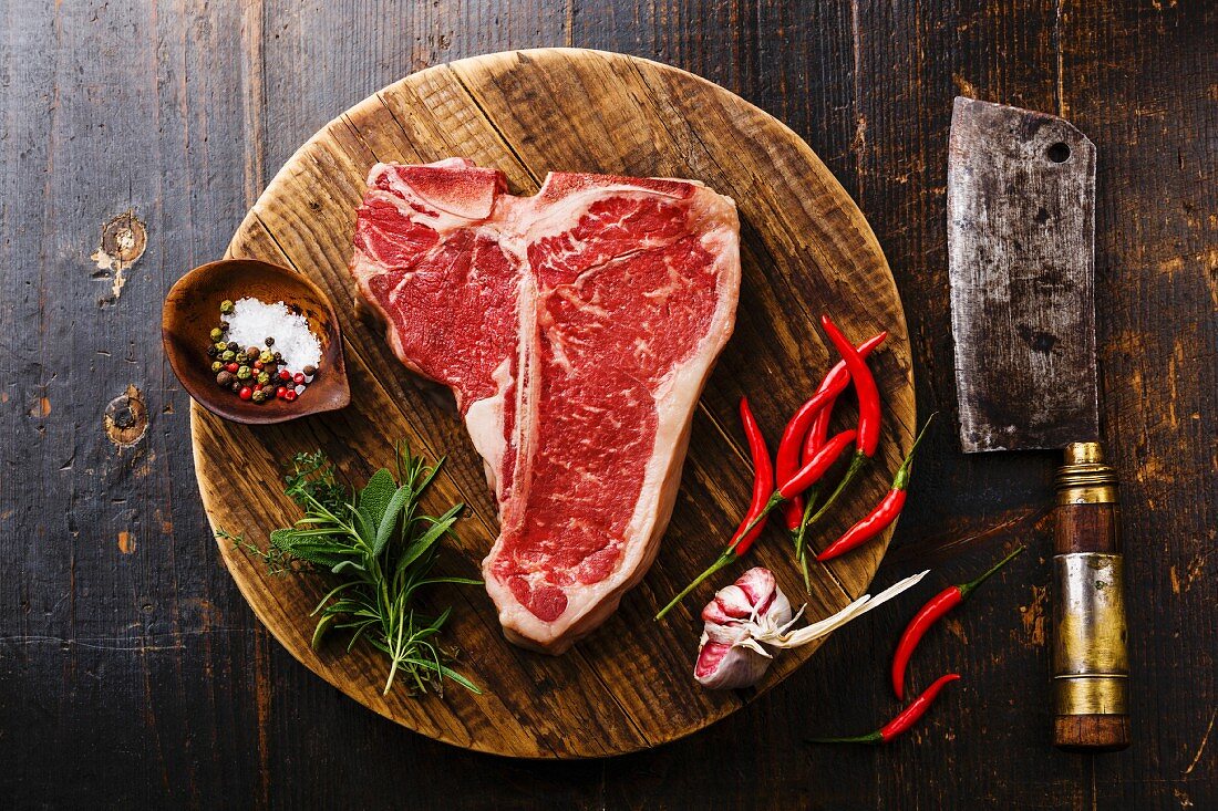 Raw fresh meat T-bone steak, seasoning and Butcher cleaver on chopping cutting board on wooden background