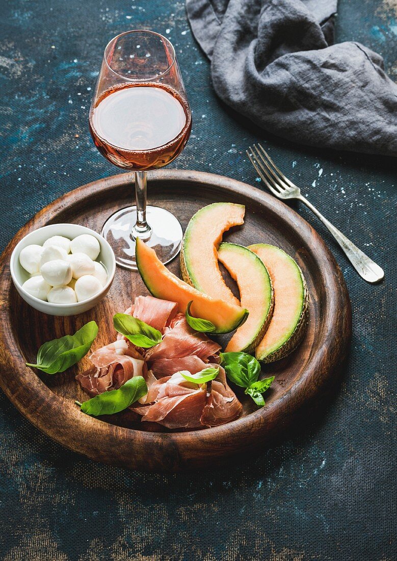 Mozzarella in a bowl, Parma ham, cantloupe melon and a glass of wine on a wooden tray