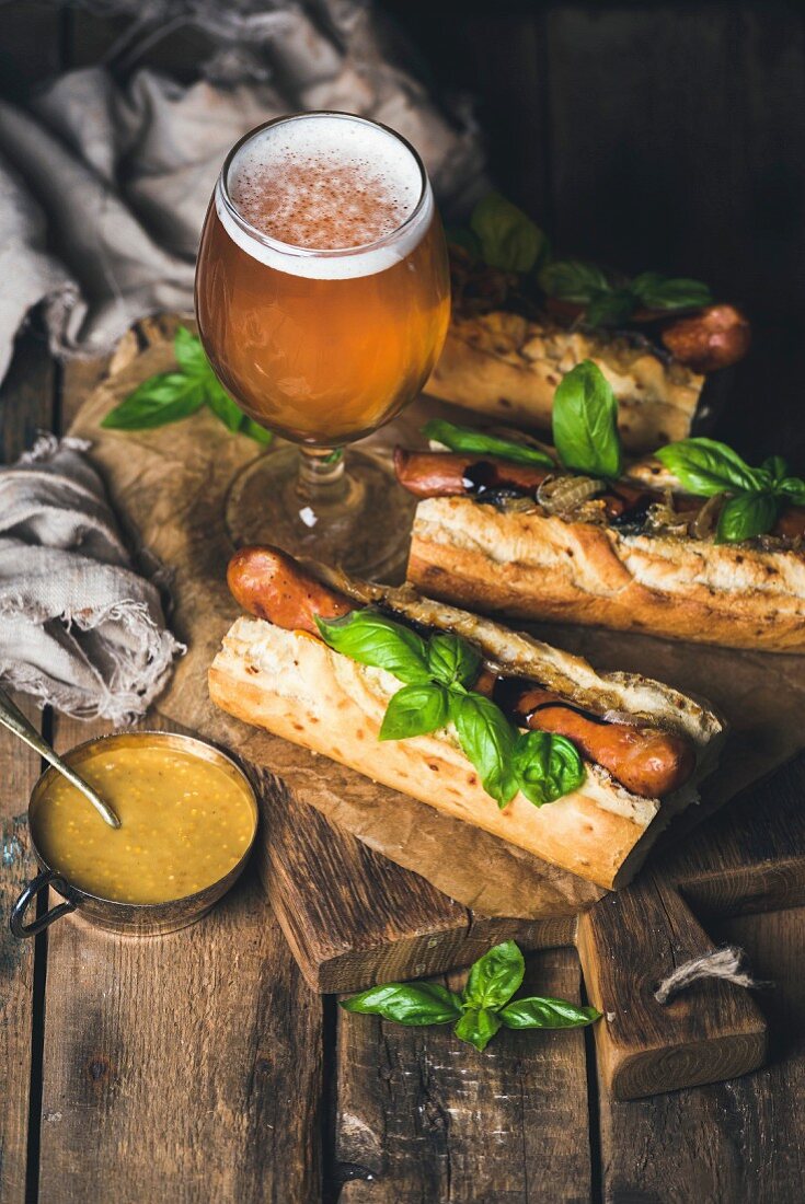 Glass of wheat unfiltered beer and grilled sausage dogs in baguette with mustard, caramelised onion and herbs