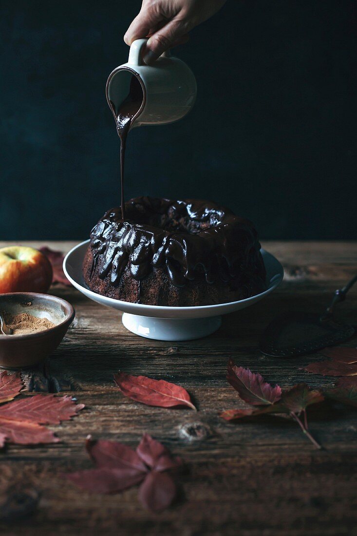 Chocolate ganache being poured onto a carob and apple Bundt cake on a cake stand