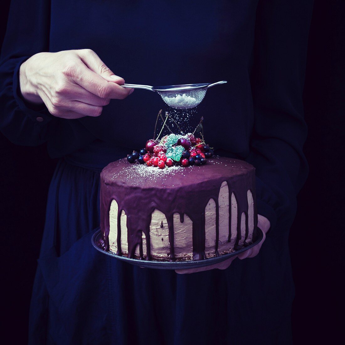 A woman's hand dusting icing sugar over a vegan chocolate cake decorated with fruits