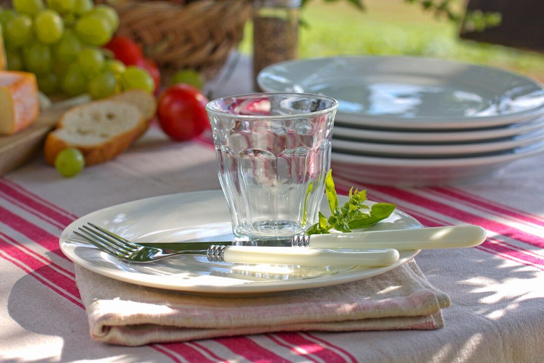 Place setting next to stacked white plates on striped tablecloth outdoors