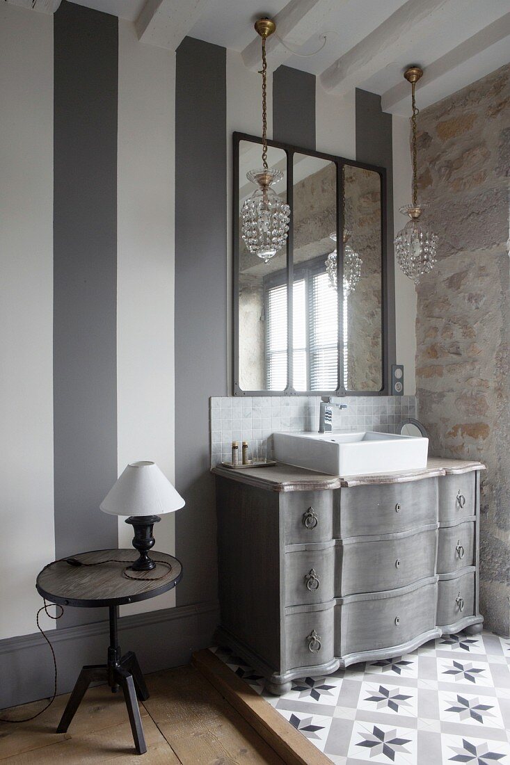 Lamp on bedside table next to sink on antique Gustavian-style chest of drawers below wall-mounted mirror