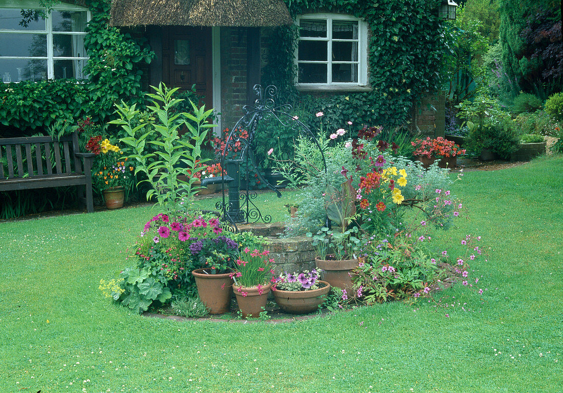 Kleiner Brunnen mit Schwengelpumpe und Töpfe mit Sommerblumen vor bewachsener Hauswand