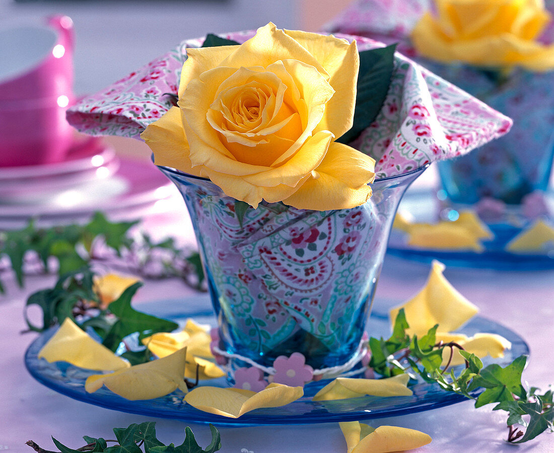 Blüte von Rosa (Rose, gelb) in kleiner blauer Vase, gemusterte Serviette