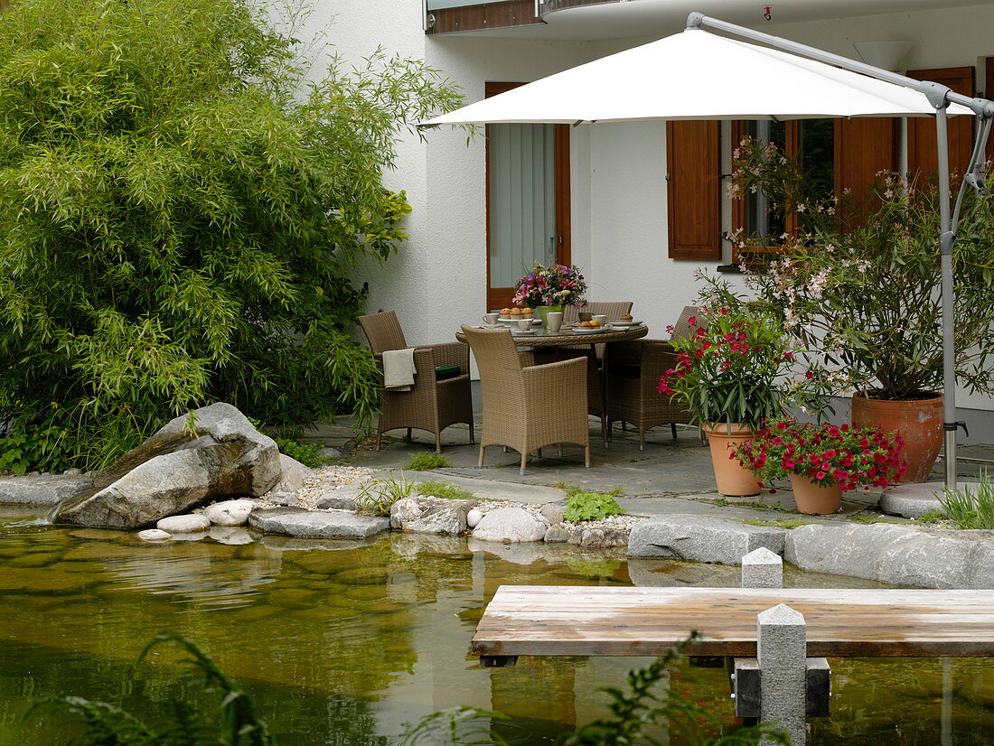 Blick vom Schwimmteich mit Holzsteg auf Terrasse mit Korbmöbeln