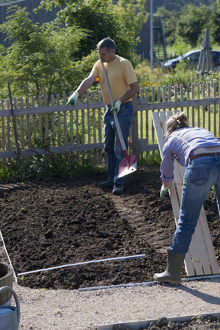 Frau legt Holzrost als Weg in fertig vorbereitetes Beet, Mann mit Schaufel