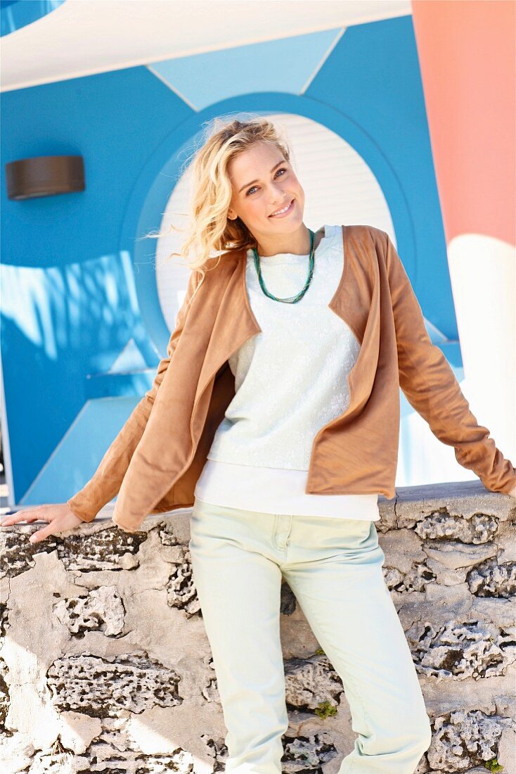 Blonde woman wearing white jumper, brown velour cardigan and mint-green jeans standing in front of wall