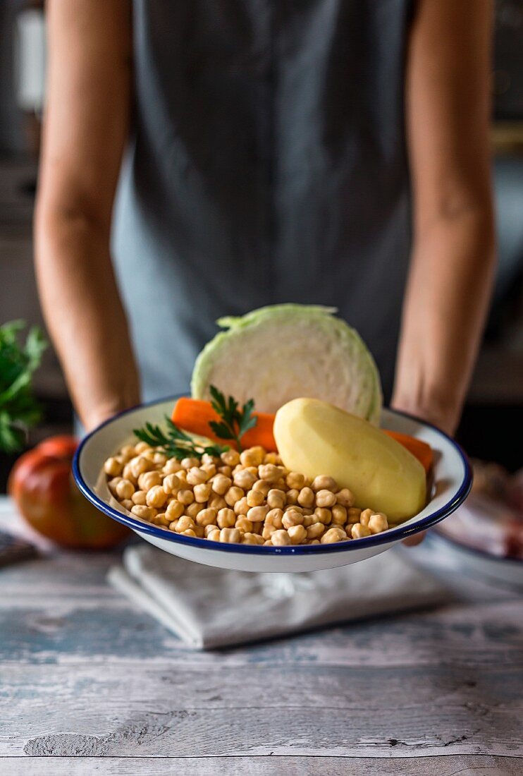 A woman holding a bowl of chickpeas, potatos, carrots and cabbage (ingredients for Cocido madrileno - a Spanish stew)