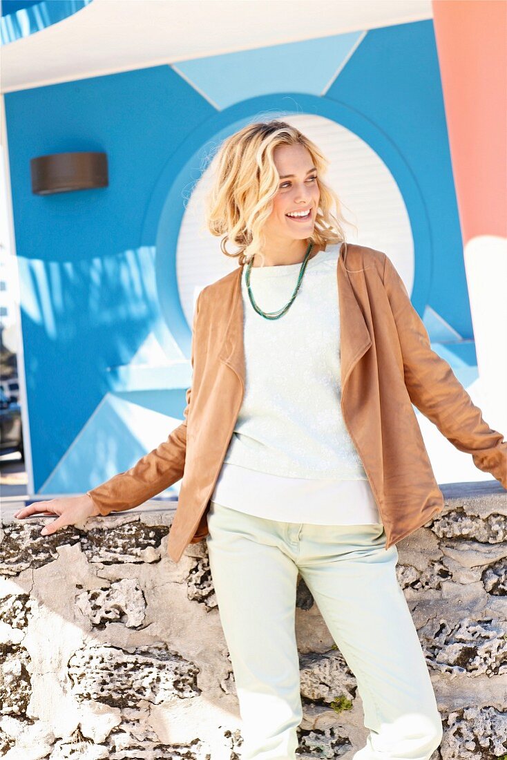 Blonde woman wearing white jumper, brown velour cardigan and mint-green jeans standing in front of wall