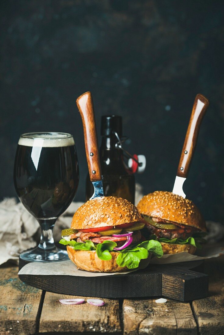 Selbst gemachte Rinderburger mit knusprigem Speck und frischem Gemüse auf Holzbrett und dunkles Bier