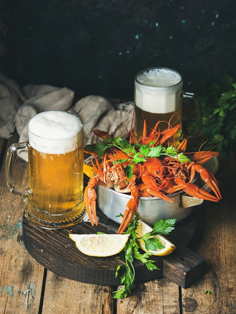 Two pints of wheat beer and boiled crayfish with lemon and parsley on a round, dark wooden serving board