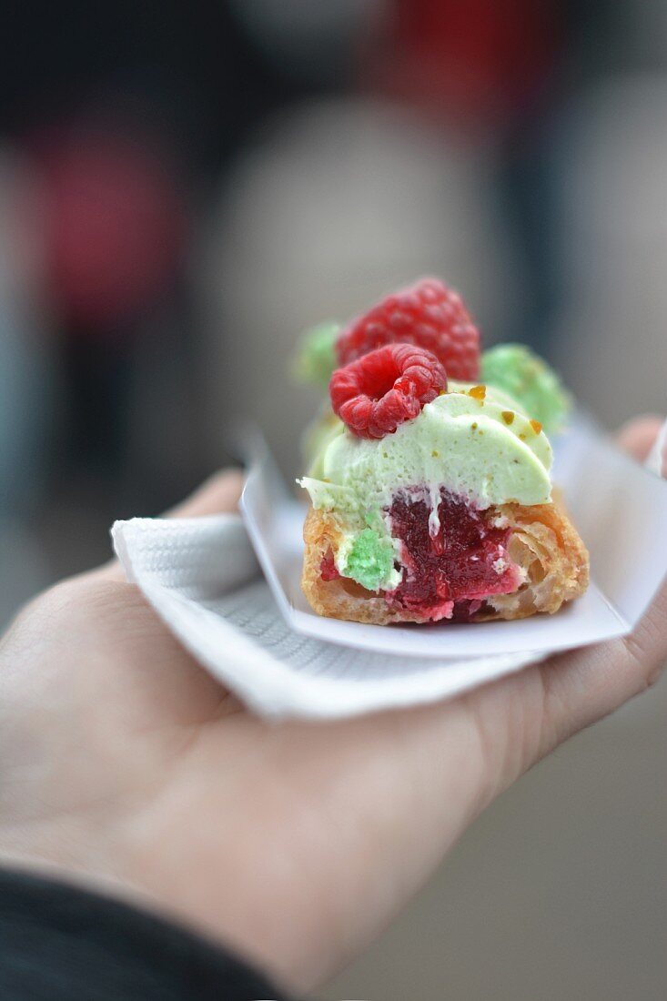 A hand holding a raspberry and pistachio eclair