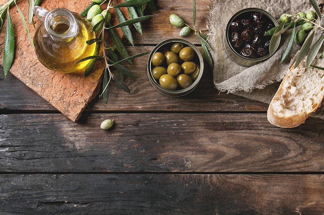 Green and black olives in tin cans with fresh bread and olives branch on sackcloth, bottle of olive oil on clay board