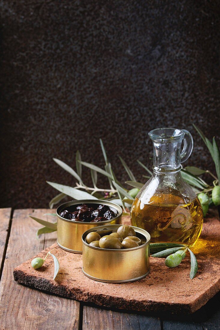 Green and black olives in tin cans, young olives branch and bottle of olive oil on clay board