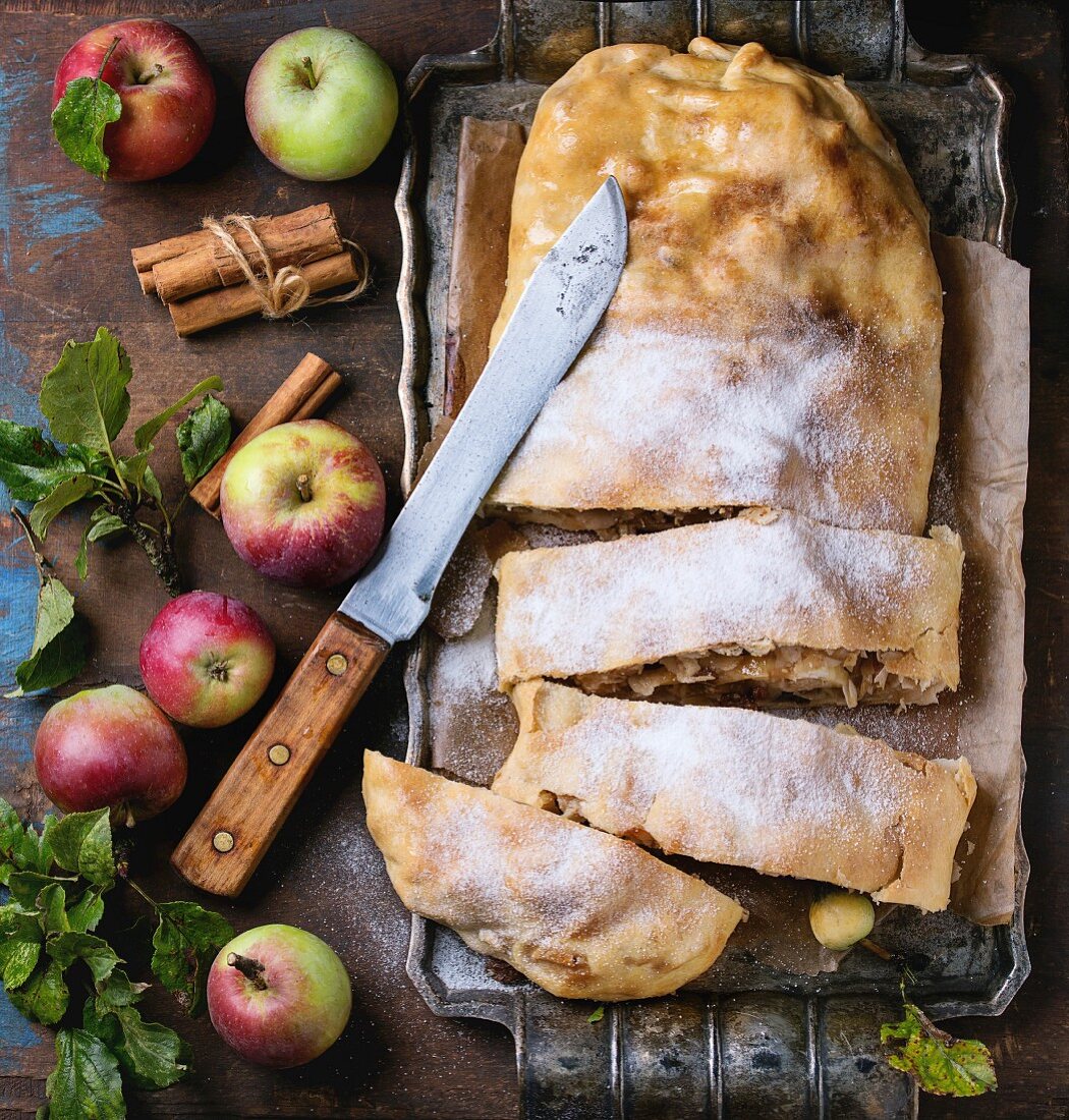 Sliced homemade apple strudel served with fresh apples with leaves, cinnamon sticks and sugar powder