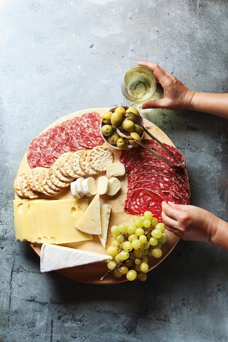 Cheese board with assorted sausages and salami, fresh fruit and various cheeses, female hand reaching salami from a platter