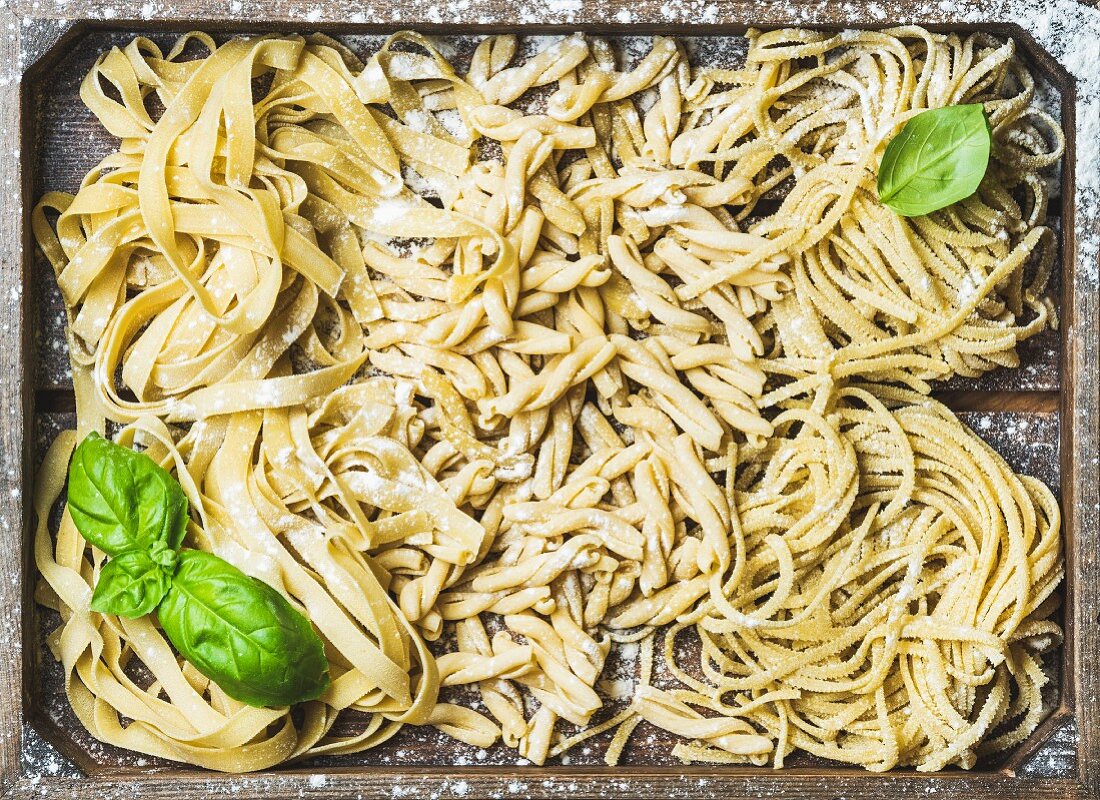 Various homemade fresh uncooked Italian pasta with flour and green basil leaves in wooden tray