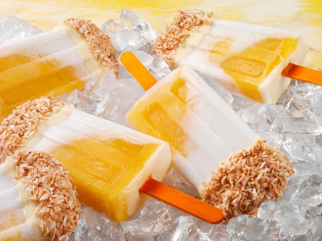 Mango-Kokos-Eis am Stiel mit weisser Schokolade und Kokosraspeln auf Eiswürfeln (Close Up)