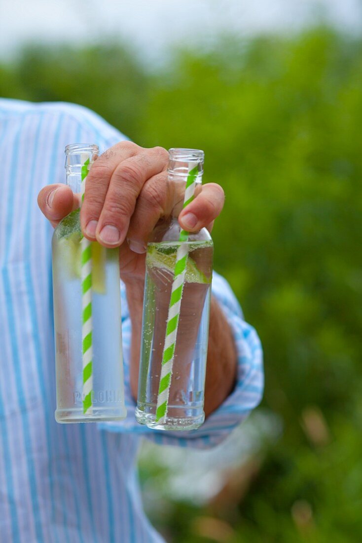 A man holding two virgin mojitos in glass bottles with straws