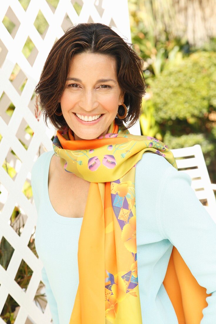 A brunette woman wearing a light blue top, hoop earrings and a patterned scarf around her neck