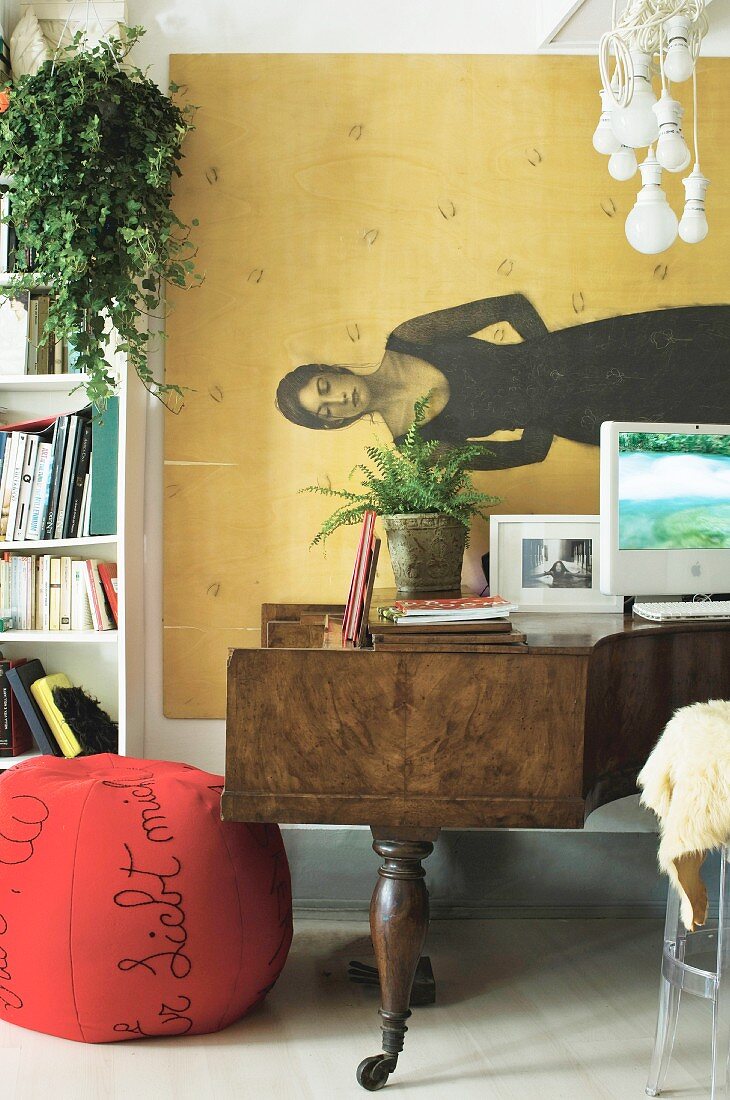 Red pouffe and computer on top of grand piano in front of painting