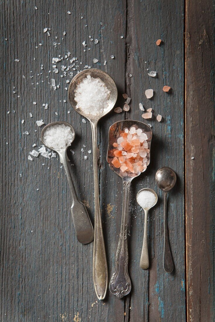 Five vintage mini spoons four filled with different types of salt and one turned over on agrey rustic wooden surface