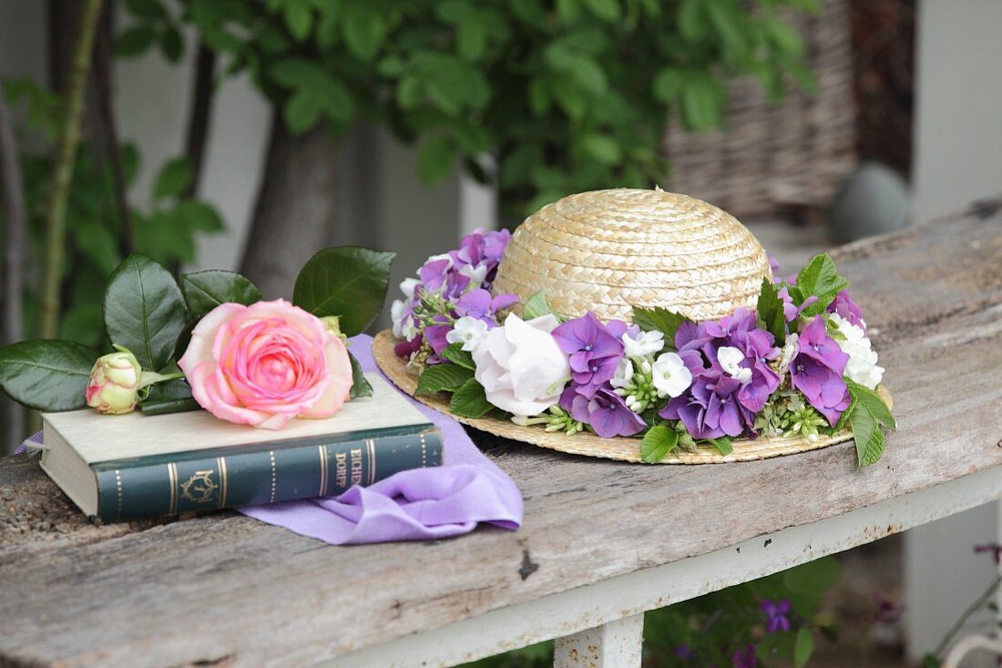 Straw hat with wreath of flowers next to rose on book in garden